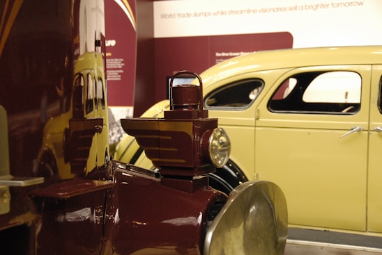 A vintage car is displayed, featuring a classic yellow body with black trim. In the foreground, a maroon antique vehicle with a lantern-style headlight is positioned, reflecting the yellow car on its polished surface. Both vehicles are highlighted by museum exhibition lighting, and descriptive text about streamlining and visionaries is visible in the background on a maroon panel.