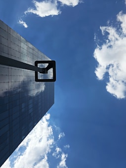 A modern skyscraper with a reflective glass facade stretches upwards against a vibrant blue sky. White fluffy clouds are scattered across the sky, some reflecting off the building's surface. A metal structure extends horizontally from the building's side, adding an element of architecture.