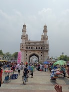 A bustling marketplace in front of a large historic monument with four minarets. Numerous people are walking around, and vendors have set up stalls selling various items including colorful garments and street food. The sky is overcast, adding a slightly subdued tone to the scene.