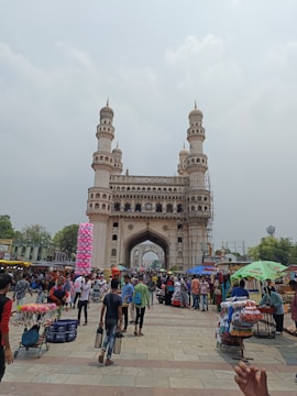 A bustling marketplace in front of a large historic monument with four minarets. Numerous people are walking around, and vendors have set up stalls selling various items including colorful garments and street food. The sky is overcast, adding a slightly subdued tone to the scene.