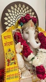A statue of Lord Ganesha adorned with intricate gold ornaments and multiple garlands of red and white flowers. A decorative backdrop features a pattern resembling lotus petals. A yellow banner with red text in a local script is draped across, containing illustrations and repeated phrases.