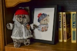 A plush toy wearing a red beanie and a gray jacket with a Saturn patch stands on a wooden bookshelf next to a framed cross-stitch of a similar bear wearing a blue and red striped hat. Several books with titles in Cyrillic script are lined up together.
