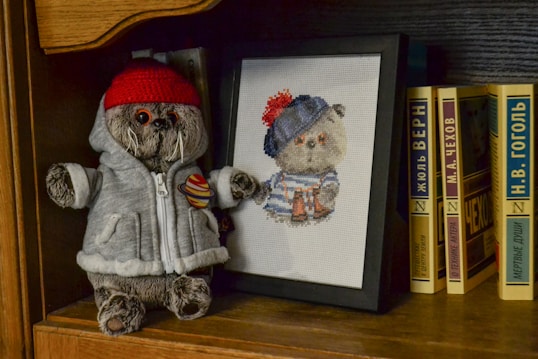 A plush toy wearing a red beanie and a gray jacket with a Saturn patch stands on a wooden bookshelf next to a framed cross-stitch of a similar bear wearing a blue and red striped hat. Several books with titles in Cyrillic script are lined up together.