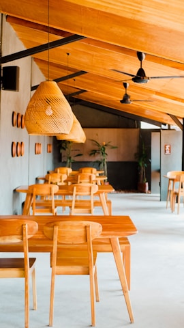 A modern cafe interior featuring wooden furniture, with several rectangular tables and matching chairs arranged neatly. The ceiling is wooden, adorned with wicker pendant lights and black ceiling fans. Plant decor is visible in the background, adding a touch of greenery.