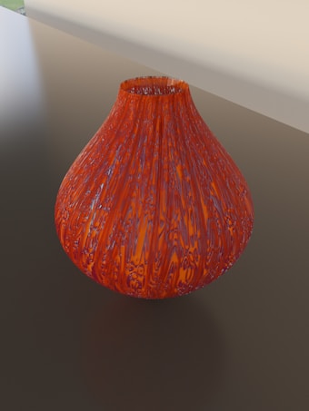 A round, intricately patterned glass vase with a reddish-orange hue sits on a reflective surface. The vase features swirling patterns that give it a dynamic and textured appearance, while the background is neutral and slightly blurred.
