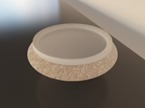 A circular object, likely a bowl or a decorative piece, with a textured surface and a smooth, recessed center. The surface features intricate, swirling patterns in gold and beige tones. The object rests on a dark, glossy surface, with a softly lit background creating a subtle gradient of light.