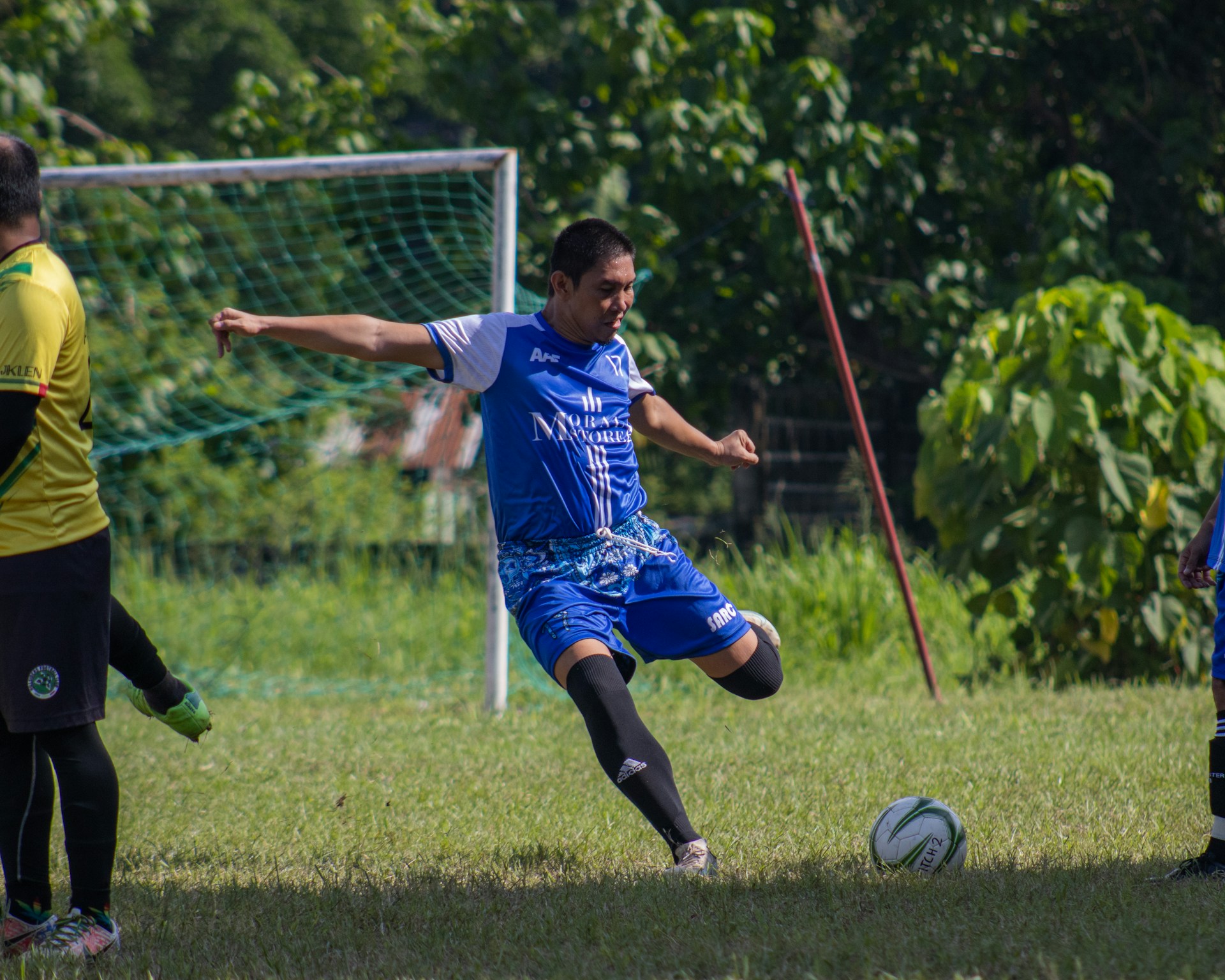 Action shot of a player wearing a bright blue and yellow football jersey, mid-kick on a sunny pitch with cheering fans in the stands.