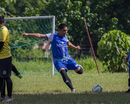 A person wearing a blue soccer uniform is mid-kick, playing on a grassy field with a goalpost in the background. Another person in a yellow jersey is visible on the left edge of the image. The scene is surrounded by lush greenery and sunlight is illuminating the players and field.
