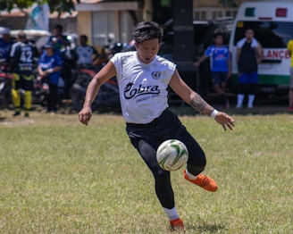 A soccer player is seen in mid-action on a grassy field, intently focusing on kicking the soccer ball with his right foot. He is wearing an orange pair of soccer cleats, black shorts, and a white jersey with visible text on it. In the background, several other people are standing and watching, and there is an ambulance and a building visible.