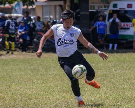 A soccer player is seen in mid-action on a grassy field, intently focusing on kicking the soccer ball with his right foot. He is wearing an orange pair of soccer cleats, black shorts, and a white jersey with visible text on it. In the background, several other people are standing and watching, and there is an ambulance and a building visible.