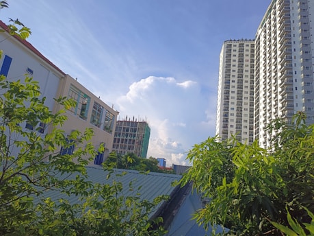 Tall modern apartment buildings and an under-construction structure rise against a clear blue sky, with vibrant green foliage from nearby trees framing the scene. The view includes several buildings with windows and balconies, indicating a residential or urban area.