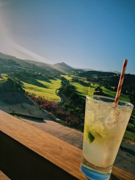 A refreshing cocktail sits on a wooden railing overlooking a scenic golf course with lush green fairways. The sun is setting in the background, casting a warm glow over the landscape.