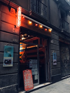 A narrow street scene featuring a bar with a neon sign, 'The Box', and a humorous sign advertising hot dogs. The entrance shows vibrant geometric patterns with warm, inviting lighting inside. The closed shutter of an adjacent establishment and images of food on the walls add to the urban atmosphere.