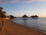 A serene beach scene at sunset with calm waters and a sandy shore. Palm trees line the left side, leading to a pier jutting into the water with traditional-style thatched pavilions on stilts.