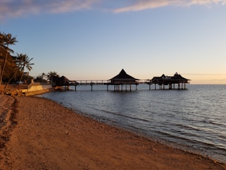 A serene beach scene at sunset with calm waters and a sandy shore. Palm trees line the left side, leading to a pier jutting into the water with traditional-style thatched pavilions on stilts.