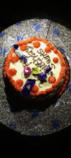 A round cake decorated with orange and white icing, featuring chocolate pieces on top and celebratory text written in brown icing. The cake is set on a floral-patterned surface.