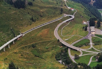 An aerial view of a winding mountain road, featuring multiple turns and bridges that navigate through a lush, green landscape. Several cars travel along the road, which cuts through the hilly terrain. The landscape is dotted with patches of trees and a few buildings near the intersection of the roads.