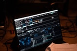 A computer screen displays video editing software with multiple timelines and controls. Various clips and tracks are arranged in a grid layout on the interface. Below the screen, some papers with printed text are partially visible, indicating a workspace environment.