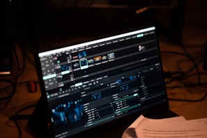 A computer screen displays video editing software with multiple timelines and controls. Various clips and tracks are arranged in a grid layout on the interface. Below the screen, some papers with printed text are partially visible, indicating a workspace environment.