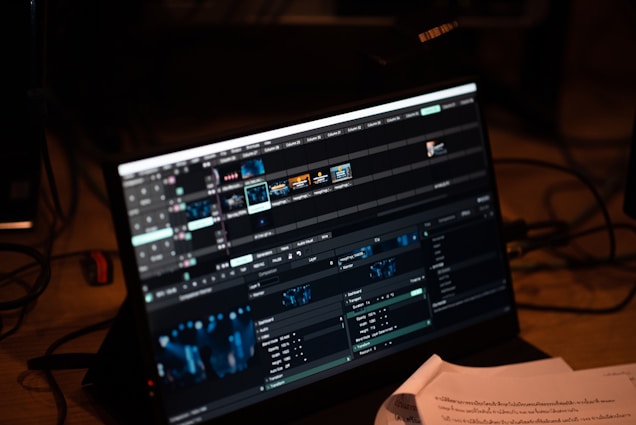 A computer screen displays video editing software with multiple timelines and controls. Various clips and tracks are arranged in a grid layout on the interface. Below the screen, some papers with printed text are partially visible, indicating a workspace environment.