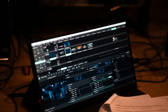 A computer screen displays video editing software with multiple timelines and controls. Various clips and tracks are arranged in a grid layout on the interface. Below the screen, some papers with printed text are partially visible, indicating a workspace environment.