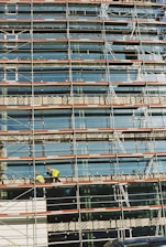A building under construction is covered with scaffolding, featuring glass panels on the facade. Two construction workers in bright safety gear are visible on the scaffold, indicating activity and progress.
