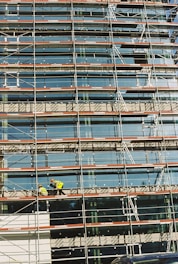A building under construction is covered with scaffolding, featuring glass panels on the facade. Two construction workers in bright safety gear are visible on the scaffold, indicating activity and progress.
