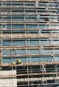 A building under construction is covered with scaffolding, featuring glass panels on the facade. Two construction workers in bright safety gear are visible on the scaffold, indicating activity and progress.