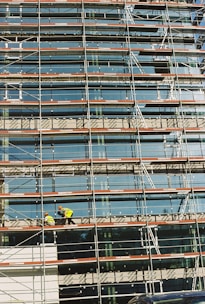 A building under construction is covered with scaffolding, featuring glass panels on the facade. Two construction workers in bright safety gear are visible on the scaffold, indicating activity and progress.