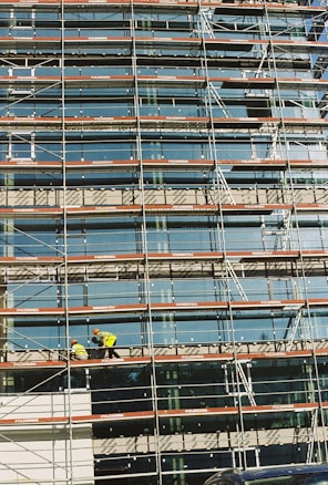 A building under construction is covered with scaffolding, featuring glass panels on the facade. Two construction workers in bright safety gear are visible on the scaffold, indicating activity and progress.