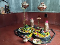 A sacred space adorned with vibrant marigold and rose flowers surrounding stone deities, including a Shivlinga. Metallic vessels hang above and a brass lamp stands lit in the center, casting a serene glow. A small altar with idols decorated with flowers is visible in the background.