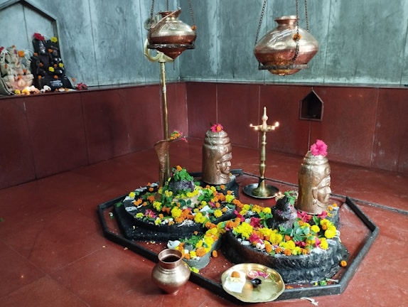 A sacred space adorned with vibrant marigold and rose flowers surrounding stone deities, including a Shivlinga. Metallic vessels hang above and a brass lamp stands lit in the center, casting a serene glow. A small altar with idols decorated with flowers is visible in the background.