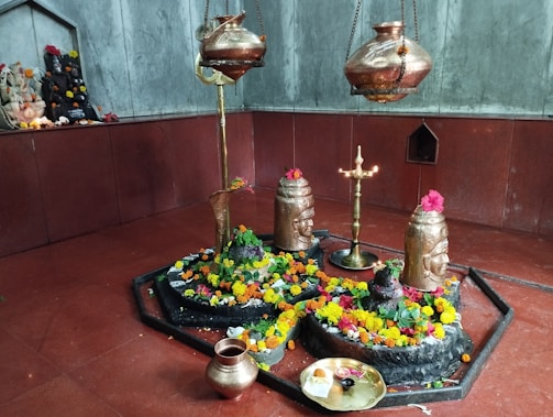 A sacred space adorned with vibrant marigold and rose flowers surrounding stone deities, including a Shivlinga. Metallic vessels hang above and a brass lamp stands lit in the center, casting a serene glow. A small altar with idols decorated with flowers is visible in the background.