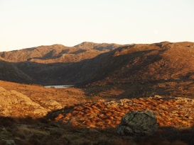 A vast mountainous landscape bathed in warm, golden sunlight. The rocky terrain features a mix of steep ridges and gently rolling hills with sparse vegetation. In the distance, a small, serene lake nestles quietly between the mountains, and the horizon is marked by rugged peaks. The colors are rich and earthy, evoking a sense of tranquility and natural beauty.