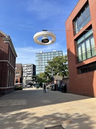 A modern urban scene with a mix of contemporary and traditional architecture. A wide pedestrian path is flanked by buildings made of red brick and glass. A unique, large overhead light fixture is suspended above the path. A few people are visible in the distance, walking near greenery and an outdoor seating area.