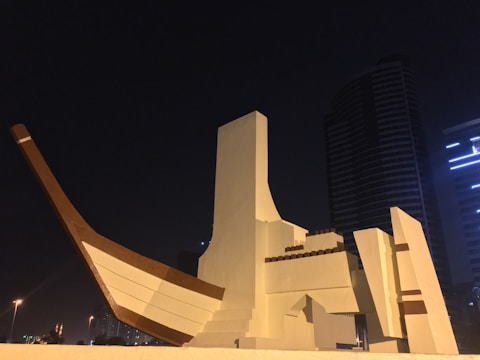 A large, abstract sculpture resembling a traditional dhow boat stands prominently against a dark, night sky. The structure is made of geometric shapes, with a combination of brown and white elements. In the background, modern high-rise buildings with illuminated windows add an urban contrast.