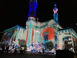 An illuminated building with intricate architectural details and colorful projections. The structure features tall minarets and a facade adorned with large circular patterns, vivid geometric designs, and abstract art. The vibrant colors create a dynamic and artistic atmosphere.