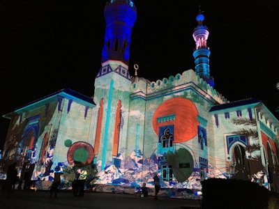 An illuminated building with intricate architectural details and colorful projections. The structure features tall minarets and a facade adorned with large circular patterns, vivid geometric designs, and abstract art. The vibrant colors create a dynamic and artistic atmosphere.