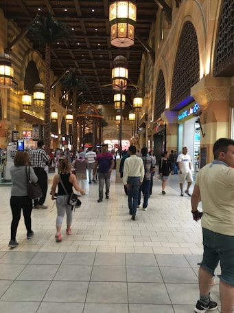 A bustling indoor shopping mall hallway with people walking in various directions. The ceiling features hanging ornate lanterns, and the architecture has an Arabesque style with arched windows and decorative elements. Shops line the sides, displaying bright signage and offers.