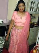 A woman wearing a traditional pink outfit stands in a kitchen. The outfit consists of a decorated blouse and a long, embellished skirt with a matching sheer dupatta. The woman is adorned with jewelry, including earrings and a necklace. Behind her, there is a countertop with a microwave and other kitchen items.