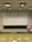 A lecture hall with green tiered seats facing a large white and black chalkboard. The room has a high ceiling with overhead lights and a ceiling-mounted projector. There are some desks and chairs in the front of the class along with a wooden podium. A fire extinguisher is visible on the wall, along with some cabinets and a sink on the right side.