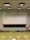 A lecture hall with green tiered seats facing a large white and black chalkboard. The room has a high ceiling with overhead lights and a ceiling-mounted projector. There are some desks and chairs in the front of the class along with a wooden podium. A fire extinguisher is visible on the wall, along with some cabinets and a sink on the right side.