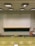 A lecture hall with green tiered seats facing a large white and black chalkboard. The room has a high ceiling with overhead lights and a ceiling-mounted projector. There are some desks and chairs in the front of the class along with a wooden podium. A fire extinguisher is visible on the wall, along with some cabinets and a sink on the right side.