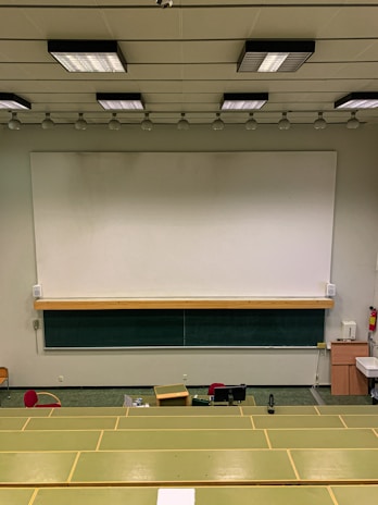 A lecture hall with green tiered seats facing a large white and black chalkboard. The room has a high ceiling with overhead lights and a ceiling-mounted projector. There are some desks and chairs in the front of the class along with a wooden podium. A fire extinguisher is visible on the wall, along with some cabinets and a sink on the right side.