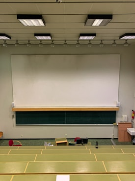 A lecture hall with green tiered seats facing a large white and black chalkboard. The room has a high ceiling with overhead lights and a ceiling-mounted projector. There are some desks and chairs in the front of the class along with a wooden podium. A fire extinguisher is visible on the wall, along with some cabinets and a sink on the right side.