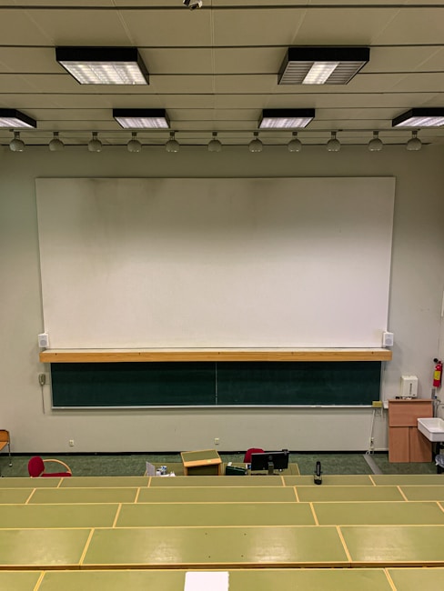 A lecture hall with green tiered seats facing a large white and black chalkboard. The room has a high ceiling with overhead lights and a ceiling-mounted projector. There are some desks and chairs in the front of the class along with a wooden podium. A fire extinguisher is visible on the wall, along with some cabinets and a sink on the right side.