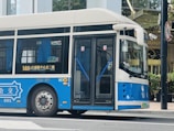 A blue and white public bus is parked on a street with glass doors and windows, displaying route information in Chinese. The bus is situated in an urban area with a hotel in the background and a road sign nearby.