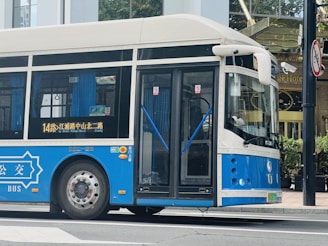 A blue and white public bus is parked on a street with glass doors and windows, displaying route information in Chinese. The bus is situated in an urban area with a hotel in the background and a road sign nearby.