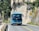 A blue tour bus is traveling on a winding mountain road with rocky cliffs on one side. Tall green trees and shrubs are visible alongside the road. There are road signs indicating a sharp turn ahead.