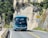 A blue tour bus is traveling on a winding mountain road with rocky cliffs on one side. Tall green trees and shrubs are visible alongside the road. There are road signs indicating a sharp turn ahead.